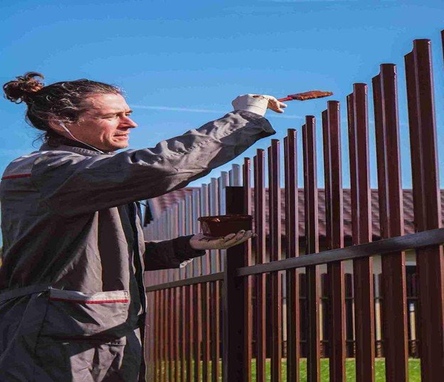 A worker painting a metal fence with a brush while holding a container of paint outdoors.